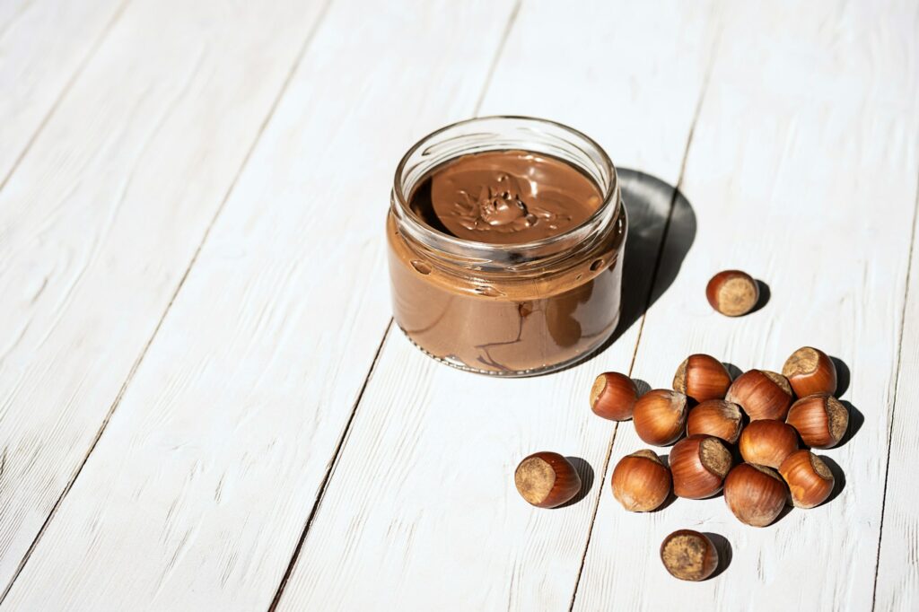 Jar of Chocolate Hazelnut Spread, Bunch of Hazelnuts on White Wooden Table.